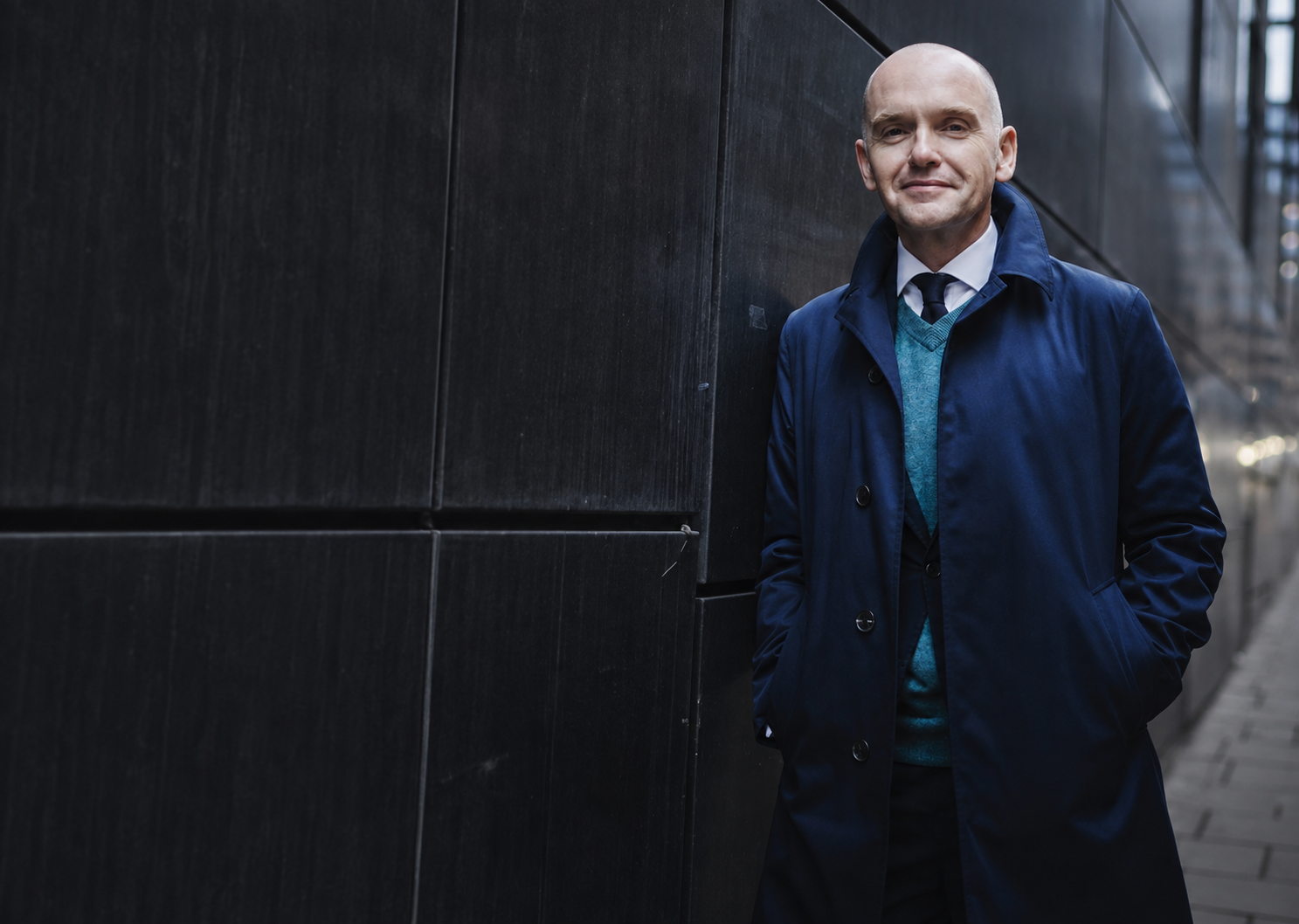 Bob Kitchen standing beside a dark stone wall in a blue coat